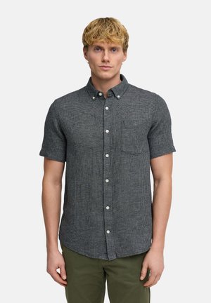 Nuori mies, jolla on vaaleat hiukset, pukeutuneena tummanharmaaseen lyhythihaiseen nappipaitaan ja oliivinvihreisiin housuihin, seisoo vaaleaa taustaa vasten.