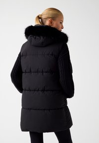 Juodas pūkuotas liemenė su dirbtiniu kailiu, su raukšlėmis megztais rankovėmis ir siuvinėtu tekstūru, kelio ilgio dizainas, su užtrauktukais kaip detalėmis.