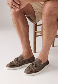Brown suede slip-on shoes with woven accents and decorative tassels, featuring a white rubber sole and a low profile design.
