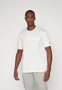 Camiseta blanca de manga corta con un bolsillo en el pecho confeccionado. La tela es suave y tiene un corte relajado. Se lleva con pantalones de chándal grises.