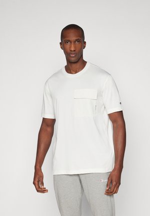 Camiseta blanca de manga corta con un bolsillo en el pecho confeccionado. La tela es suave y tiene un corte relajado. Se lleva con pantalones de chándal grises.