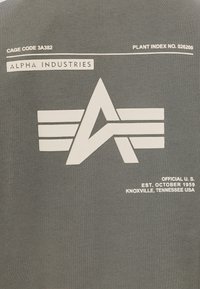 T-shirt grigia con grafica bianca rappresentante una "A" e un design con ali. Include dettagli testuali: "ALPHA INDUSTRIES" e informazioni di produzione.