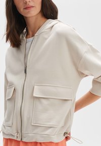 Felpa con zip di colore beige chiaro, con finitura testurizzata, dotata di due tasche frontali e di un cordino all'orlo. Tessuto in misto cotone.