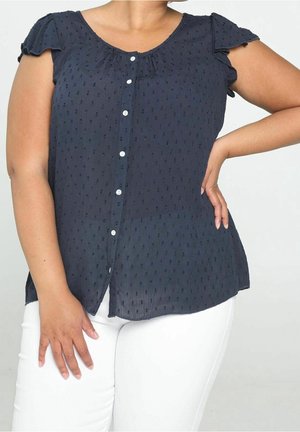 Femme portant un chemisier bleu marine à manches courtes avec des boutons blancs et des points texturés, associé à un pantalon blanc, posant avec une main sur la hanche.