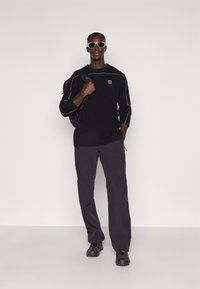 Felpa nera con cuciture bianche, logo sul petto; pantaloni grigio scuro; zaino nero; occhiali da sole; scarpe sportive nere.