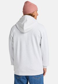 Sudadera con capucha de color gris claro, de corte holgado, con puños acanalados y detalle de costura en la parte posterior. Combinada con jeans de mezclilla azul y un gorro de punto rosa.