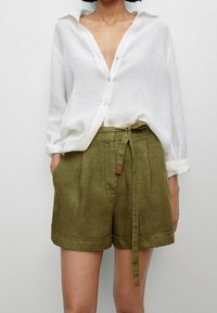 Camicia bianca in lino con maniche arrotolate abbinata a pantaloni corti verdi in lino a vita alta con cintura e orlo risvolto.