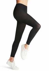 Schwarze, knöchellange Leggings aus dehnbarem Stoff mit hohem Bund und glatter, matter Textur, kombiniert mit weißen Sneakern.