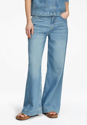 Kobieta ubrana w jasnoniebieskie jeansy o szerokich nogawkach, kurtkę dżinsową i brązowe płaskie sandały, stojąca z jedną ręką w kieszeni.