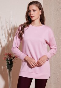 Felpa rosa chiaro a maniche lunghe con scollatura rotonda, fessure laterali all'orlo e texture morbida e liscia. Abbinata a leggings scuri.