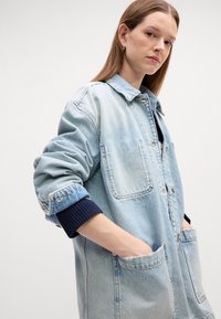 Veste en denim bleu clair avec deux poches avant, fermeture à boutons et poignets côtelés bleu marine. Coupe légèrement oversize avec une texture douce.