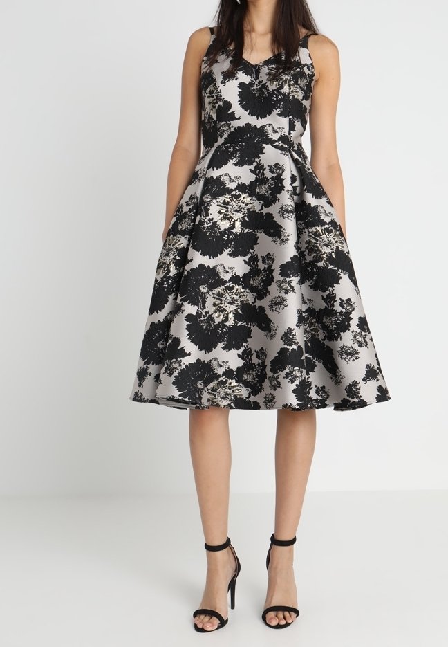 Femme portant une robe sans manches en ligne A avec de grands motifs floraux noirs sur fond gris argenté et des sandales à talons hauts noires.