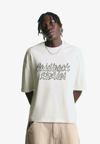 Bílé oversized tričko vyrobené z bavlny, s černým vyšívaným textem, který zní "laidback stories". Krátké rukávy a uvolněný střih.