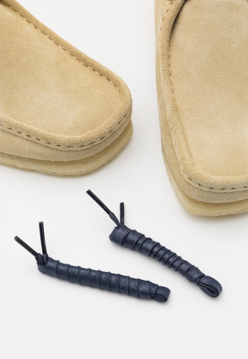 Mocassino in suede tan con bordi cuciti, abbinato a lacci intrecciati di colore blu scuro per la chiusura, dotato di suola piatta.