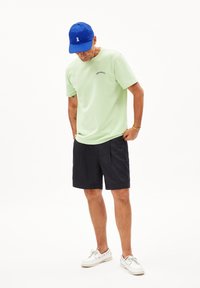  Lichtgroene T-shirt met korte mouwen en een gebogen logo, gecombineerd met donkerblauwe shorts. Een blauwe pet en witte schoenen maken de outfit compleet.