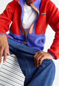 Chaqueta de forro polar roja y morada con un detalle de cremallera naranja, que cuenta con dos bolsillos frontales y un ajuste casual. Combinada con jeans de mezclilla.