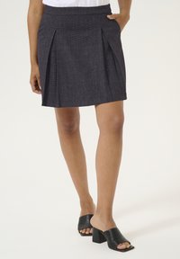 Jupe plissée gris foncé avec une finition texturée, présentant une taille ajustée et un ourlet évasé. Associée à des mules noires à bout ouvert.