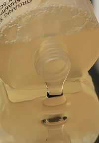 Ett ekologiskt schampoflaska med en genomskinlig gul vätska som hälls från en klar pip, reflekterande på en svart yta. Bubblor synliga.