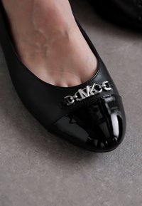 Zwarte leren platte schoen met een gepatineerde leren neuskap en een zilveren logoaccent. Glad textuur, ronde vorm, gedetailleerde gespsieraden.
