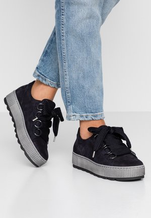 Persoon draagt een blauwe spijkerbroek en zwarte platformsneakers met dikke zwarte veters, staand op een reflecterende witte vloer.