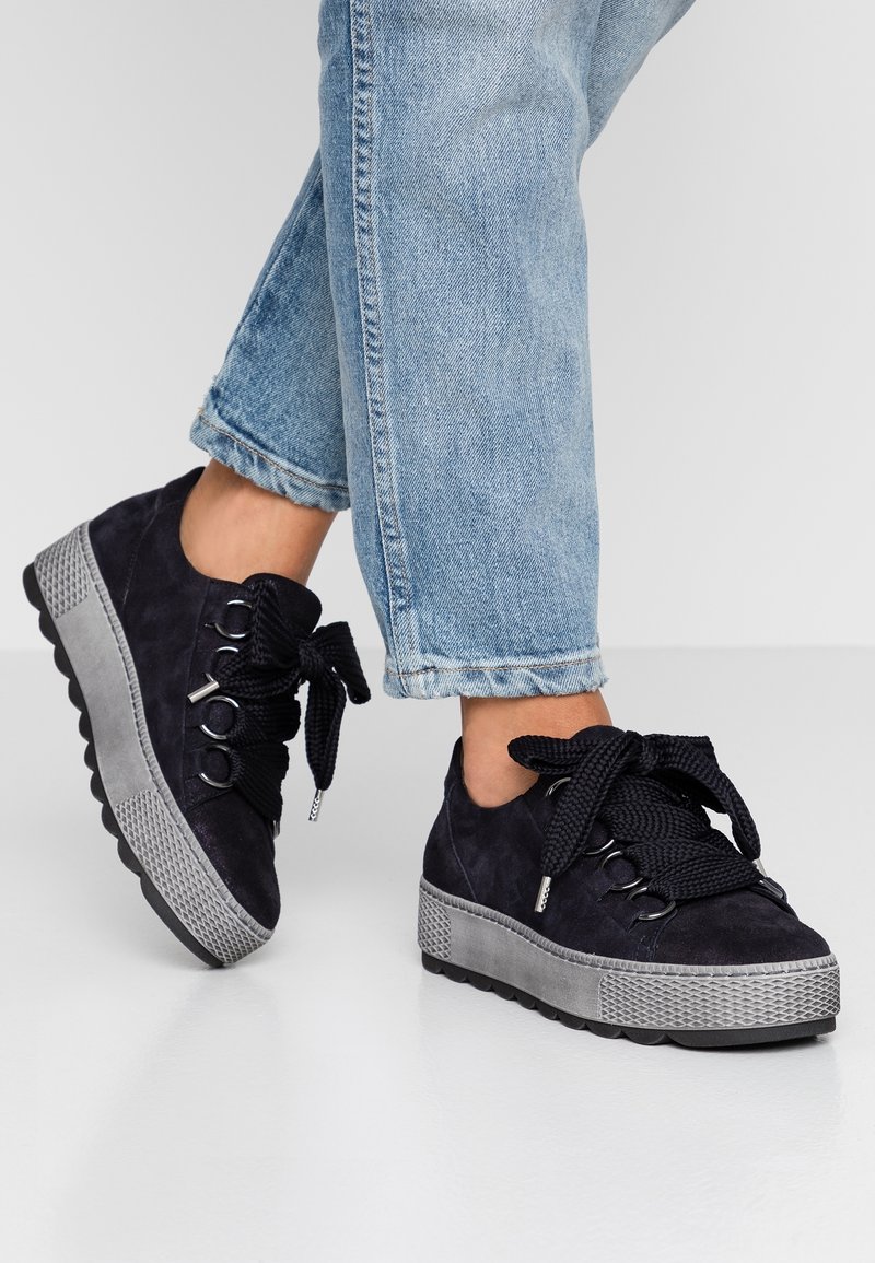 Personne portant un jean bleu et des baskets compensées noires avec de gros lacets noirs, debout sur un sol blanc réfléchissant.
