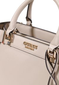 Sac à main en cuir beige avec quincaillerie dorée. Présente des poignées supérieures, une fermeture à glissière et le logo "GUESS" embossé à l'avant. Texture lisse.