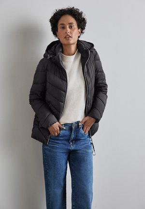 Schwarze Pufferjacke mit horizontalem Steppmuster, verstellbarer Kapuze, Reißverschlüssen und Taschen, getragen über einem hellen Pullover und blauen weiten Jeans.