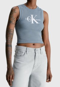Débardeur court à rayures de couleur bleu clair avec le logo blanc "Calvin Klein Jeans". Associé à un jean en denim gris clair taille haute.