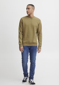 Olive green sweatshirt with a crew neck and ribbed cuffs, paired with blue jeans and black sneakers in a casual standing pose.