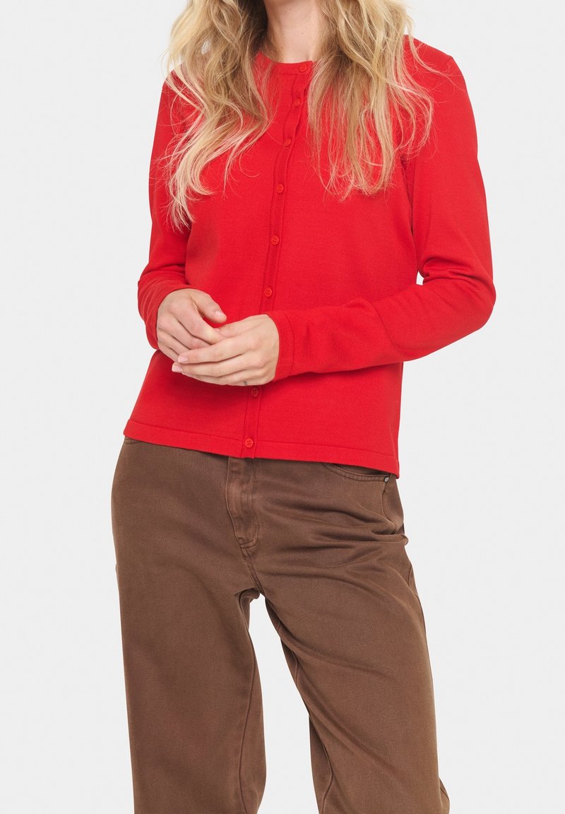 Cardigan rouge à boutons, fabriqué en tissu doux, avec des manches longues et un ourlet arrondi. Porté avec un pantalon marron, les mains jointes devant.