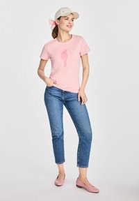 Różowa bawełniana koszulka z grafiką flaminga, zestawiona z jasnoniebieskimi dżinsami i różowymi balerinami. Całość dopełnia beżowa czapka.