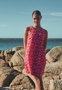 Femme portant une robe rose sans manches à motif coquillage, debout sur une rive rocheuse avec l'océan et le ciel en arrière-plan.