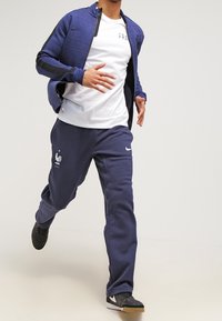 Giacca sportiva navy con finitura texturizzata, abbinata a una maglietta bianca e pantaloni jogger navy con logo di gallo e accenti Nike.