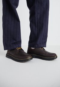 Rjave usnjene čevlje z rumenim šivom, okroglo konico in vezalkami, v kombinaciji z mornarskimi črtastimi hlačami, ki prikazujejo vsakdanji dizajn.