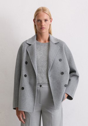 Femme aux cheveux blonds raides portant un manteau double boutonnage gris clair, un pull en tricot et un pantalon taille haute, se tenant devant un mur blanc.