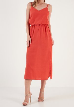 Vrouw draagt een mouwloze oranje midi-jurk met dunne bandjes, een geplooide taille, een zijsplit en beige sandalen met hoge hakken.