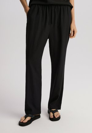 Czarne, lekkie, długie spodnie z elastycznym paskiem w talii i bocznymi kieszeniami. W połączeniu z czarnymi klapkami na gołych stopach, na neutralnym tle.