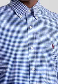 Niebiesko-biała koszula w kratę gingham z kołnierzykiem rozłożystym i białymi guzikami. Na piersi znajduje się małe czerwone haftowane logo.
