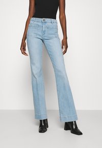 Hellblaue, ausgestellte Jeans aus Denim mit hoher Taille, zwei Gesäßtaschen und einem Knopfverschluss. Kombiniert mit schwarzen Knöchelstiefeln.