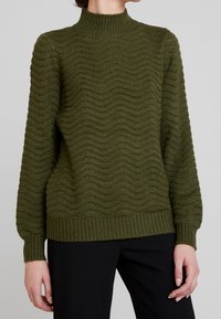 Maglione lavorato a maglia verde oliva con collo alto, polsini e corpo a coste. Presenta un motivo a trama ondulata su tutta la superficie. Indossato con pantaloni neri.
