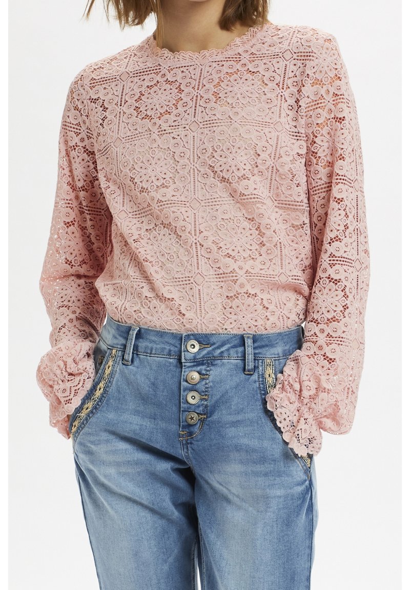 Pink lace long-sleeve top featuring a floral pattern, scalloped neckline, and textured fabric, paired with light blue denim jeans with button detail.