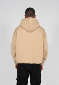 Sweat à capuche beige avec une coupe décontractée, poignets et ourlet côtelés, vu de l'arrière. Fabriqué en tissu doux, contrastant avec un pantalon noir.