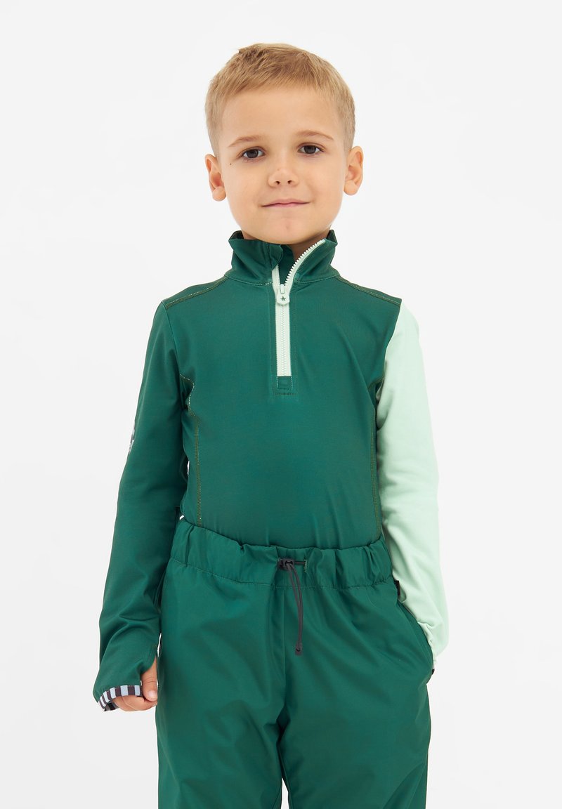Grüner Reißverschlussoberteil mit hohem Kragen und hellgrünen Ärmeln, kombiniert mit dunkelgrünen elastischen Hosen mit höherem Bund. Glattes Material, minimalistische Details.