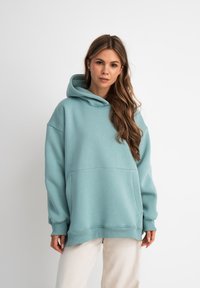 Felpa oversized color teal chiaro realizzata in tessuto morbido, con una comoda tasca frontale e cappuccio con coulisse, abbinata a pantaloni color crema.