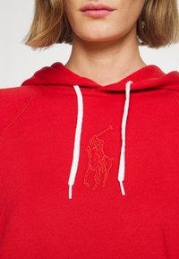 Červená mikina s kapucí vyrobená z jemné tkaniny, která má bílou šňůrku a vyšitý logem koně na prsou. Neformální design s raglánovými rukávy.