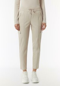 Beige cargobroek met een elastische tailleband en trekkoord, voorzien van ritssluitingen aan de voorkant en zijcargo zakken. Glad, lichtgewicht materiaal.