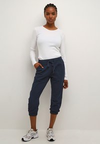 Top blanco de manga larga con cuello redondo, combinado con joggers azul marino con cordón y zapatillas deportivas grises. Tejido suave, ajuste casual.