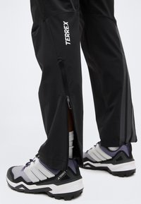 Κατώτερα πόδια που φορούν μαύρο παντελόνι TERREX με πλαϊνά φερμουάρ, σε συνδυασμό με γκρι και μαύρα παπούτσια πεζοπορίας TERREX και λευκές κάλτσες.
