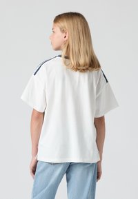 Balts īsām piedurknēm T-krekls no kokvilnas ar tumši ziliem svītrām uz pleciem. Brīvi pieguļoša silueta, skats no aizmugures, apvienots ar gaiši ziliem džinsiem.