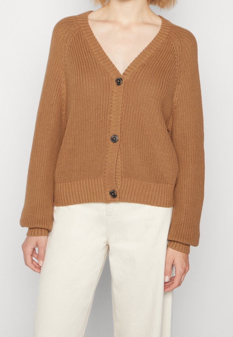 Personne portant un cardigan en maille marron avec trois boutons noirs et un pantalon blanc, debout devant un fond clair uni.